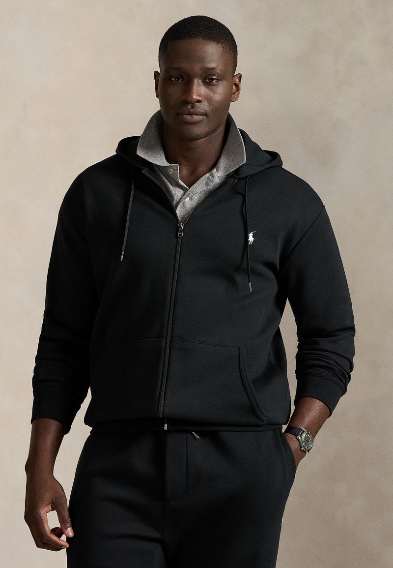 Svart zip-up hoodie i mjukt tyg, med grått innerfoder, kängurufickor och en liten vit logga på bröstet.