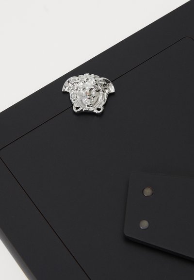 Dosar din piele neagră, decorat cu un emblemă argintie în formă de cap de Meduză. Include un capac cu clapetă și doi nituri metalice pentru un plus de detaliu.