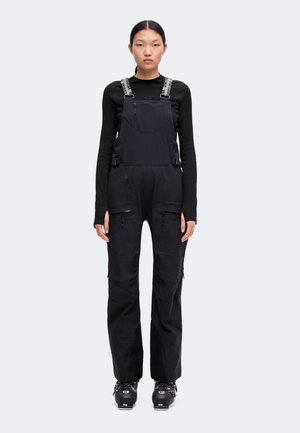 Jeune femme debout portant un t-shirt noir à manches longues, une salopette noire avec des poches zippées et des chaussures de ski noires sur un fond uni.