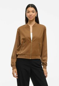 Femme aux longs cheveux noirs portant une veste bomber brune zippée, un pantalon noir et des boucles d'oreilles argentées, se tenant devant un fond blanc.