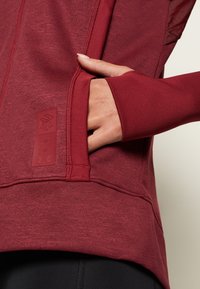 Main placée dans la poche avant d'une veste de sport bordeaux à manches longues avec trou pour le pouce sur le poignet et tissu texturé.