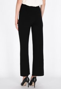 Zwarte wijde broek van achteraf gezien, gemaakt van zachte, gestructureerde stof, met een hoge taille en een gladde afwerking. Draagt zwarte hoge hakken.