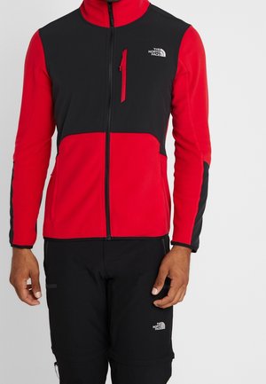 Person trägt eine rot-schwarze North Face Jacke mit Reißverschluss und schwarze North Face Hose, steht vor einem einfarbigen Hintergrund.