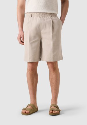 Beige knee-length shorts with elastic waistband worn by a standing person, paired with olive green double-strap sandals.