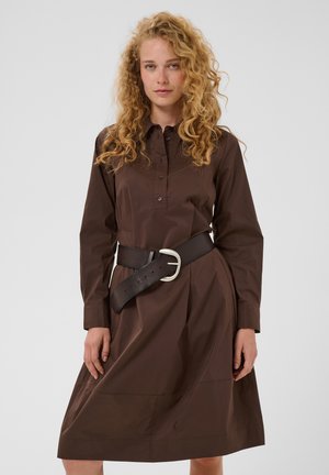 Femme aux cheveux blonds bouclés portant une robe marron mi-longue avec des manches longues et une large ceinture noire avec une boucle argentée.