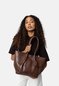 Brązowa skórzana torba typu tote z dwiema długimi uchwytami, charakteryzująca się miękką fakturą i przeszyciami. Torba jest pojemna i bez sztywnej formy.