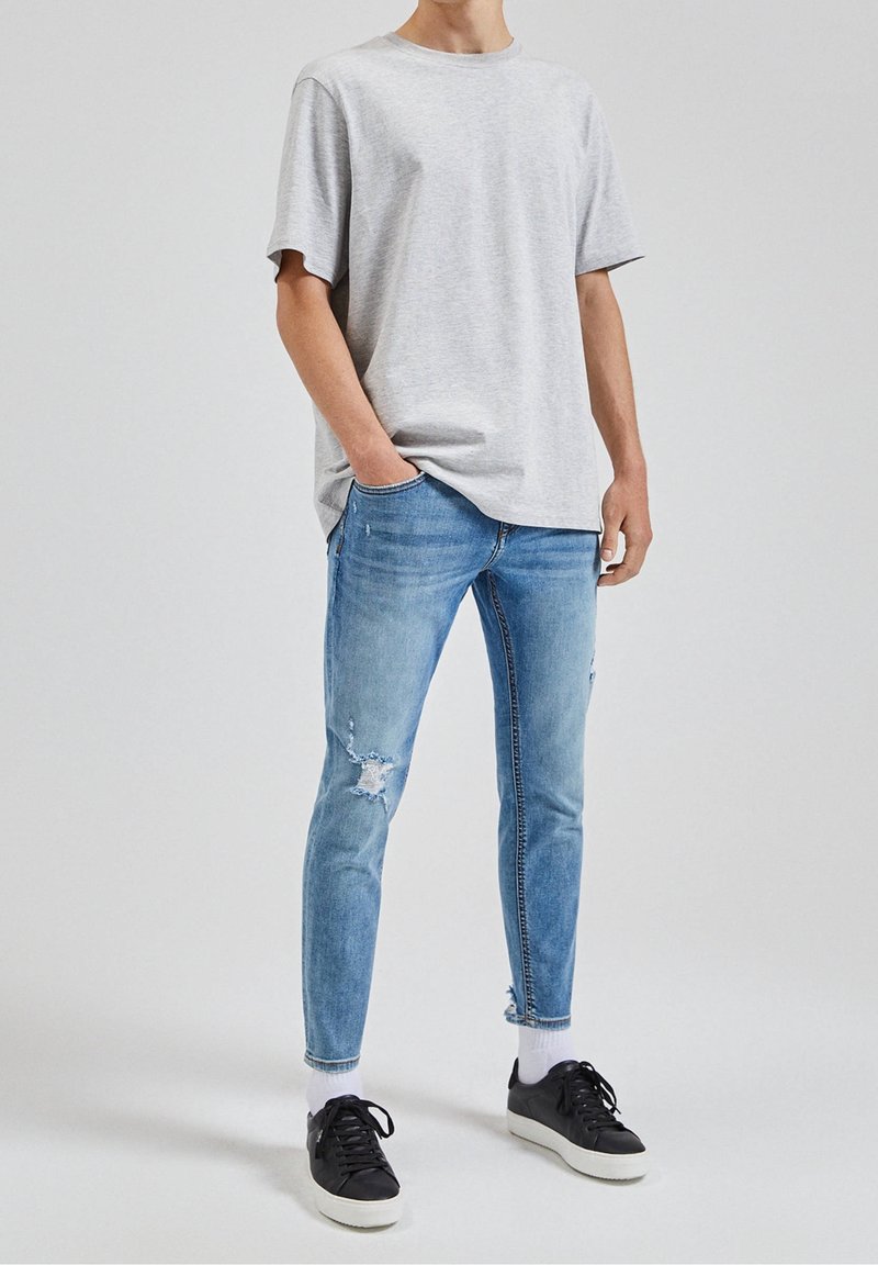 Grå bomullströja i kombination med ljusblå slitna jeans. Jeanserna har revor och en tapered passform. Svarta sneakers med vita sulor fulländar outfiten.