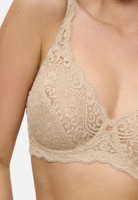 Beige pitsikoristeilla varustettu rintaliivi, jossa on kukkakuviot, henkilöllä. Näkyvissä yksityiskohtainen kirjonta kupin ja olkaimien alueella.