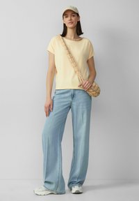 Jasnożółta bluzka z krótkim rękawem, luźne jasnoniebieskie dżinsy, stylizowane z plecioną torebką crossbody w kolorze beżowym oraz białymi sneakersami.