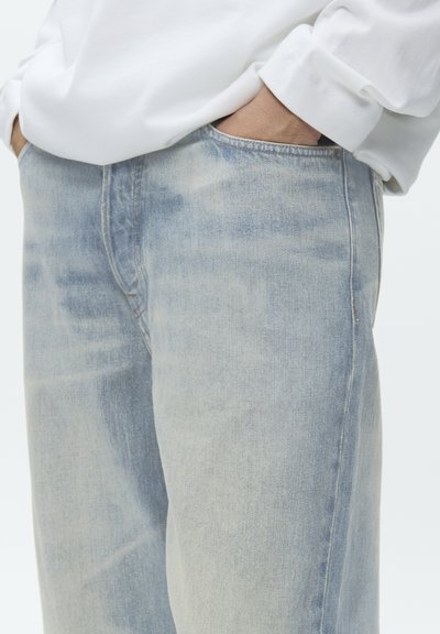 Ruky zasunuté vreckách svetlomodrých vyblednutých džínsov, ktoré sú nosené s bielou dlhorukávovou košeľou.