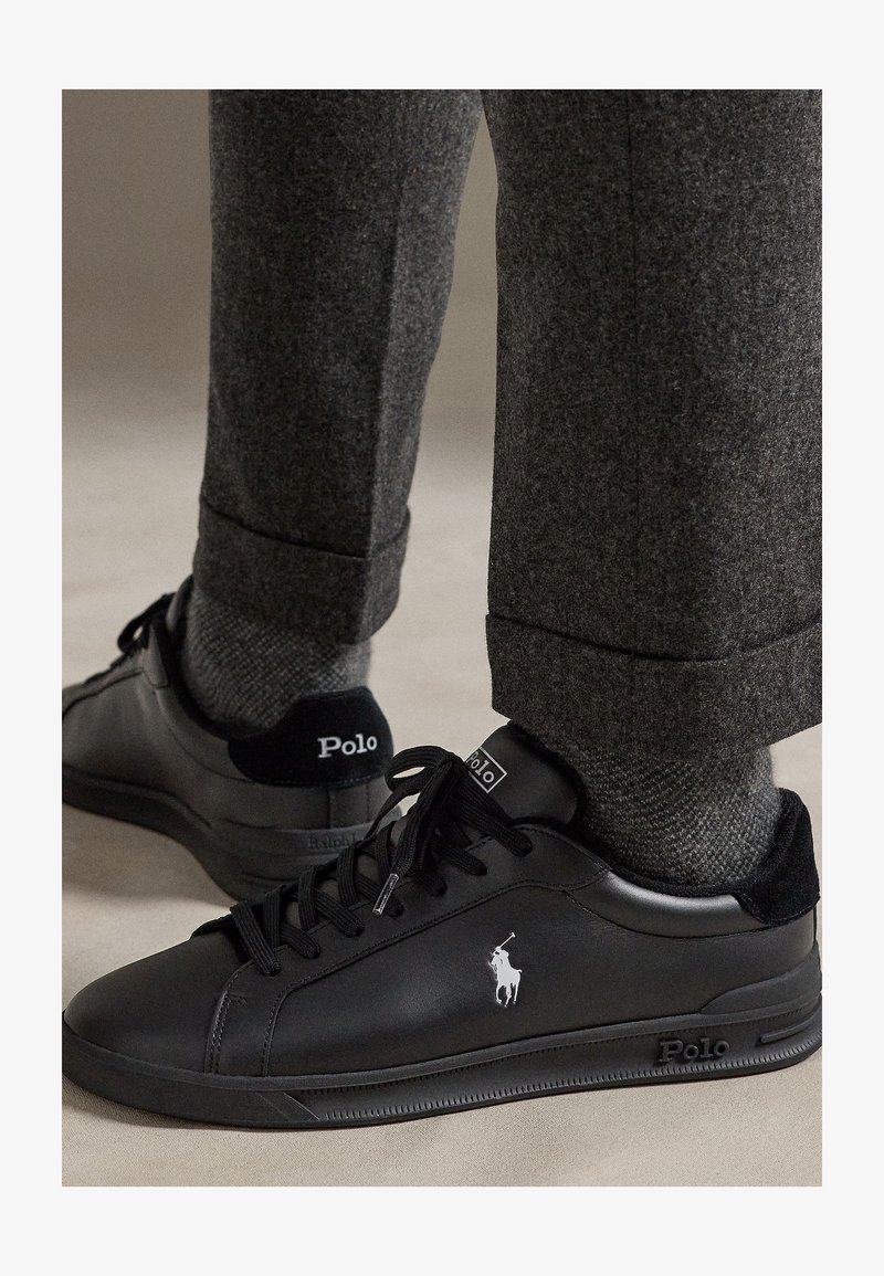 Juodi Polo Ralph Lauren sportiniai bateliai su baltu logotipu, dėvimi su pilkomis vilnonėmis kelnėmis ir derančiomis pilkomis tekstūruotomis kojinėmis ant neutralaus grindinio.