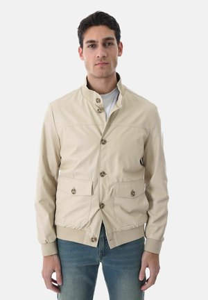 Giovane uomo che indossa una giacca beige abbottonata con tasche a patta e polsini a coste, abbinata a jeans blu, in piedi contro uno sfondo semplice.