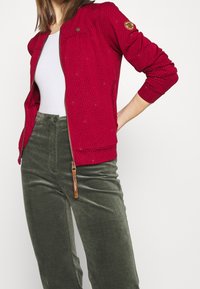 Veste bomber rouge zippée avec un motif texturé et des poignets élastiques, associée à un pantalon en velours côtelé vert olive doté d'une fermeture éclair latérale et d'une fermeture à bouton.