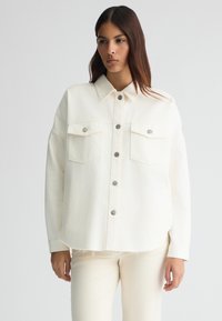 Giubbotto di jeans crema con collo a camicia, chiusura frontale a bottoni, due tasche sul petto e orlo sfilacciato. Indossato sopra pantaloni abbinati di colore chiaro.