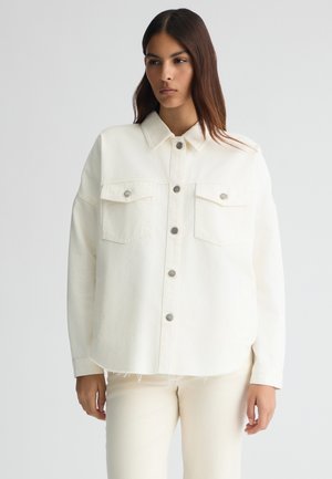 Giubbotto di jeans crema con collo a camicia, chiusura frontale a bottoni, due tasche sul petto e orlo sfilacciato. Indossato sopra pantaloni abbinati di colore chiaro.