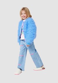 Giacca fluffy azzurro chiaro con texture arricciata, t-shirt grafica, jeans ricamati in lavaggio chiaro con testo rosa e scarpe bianche slip-on.