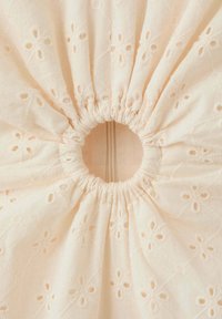 Tissu couleur crème avec broderie florale ajourée rassemblée autour d'une ouverture élastique centrale, créant un motif froncé circulaire.
