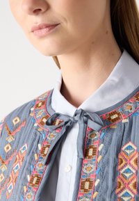 Indumento grigio ricamato con motivi intricati multicolor. Presenta una chiusura a cravatta e una blusa bianca liscia sotto.