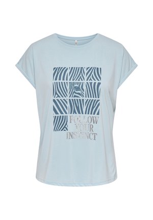 Joven mujer con camiseta gráfica azul clara con el texto "Follow Your Instinct", pantalones cortos negros con borde ondulado y sandalias negras tipo slide, de pie.