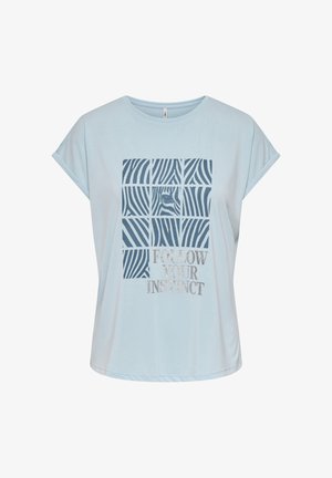 Joven mujer con camiseta gráfica azul clara con el texto "Follow Your Instinct", pantalones cortos negros con borde ondulado y sandalias negras tipo slide, de pie.
