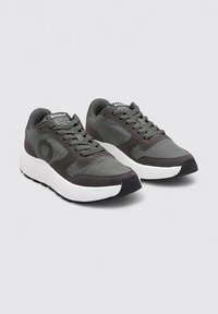 Baskets grises en matériaux mixtes avec un col rembourré, des lacets plats, une semelle intermédiaire blanche, et un détail de logo sur le côté. Semelle extérieure en caoutchouc noir.