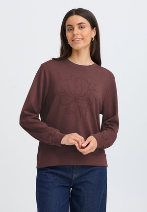 Kvinde i en brun sweatshirt med et svagt blomsterdesign og blå jeans, stående med hænder løst foldet og let smilede.