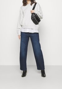 Osoba v světlém šedém svetru, bílém tričku, tmavě modrých širokých džínách, černých kotníkových botách a držící černou crossbody tašku.