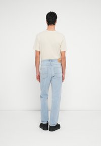 Világoskék egyenes szárú farmernadrág hátul zsebekkel és barna bőrpánttal a deréknál; bézs színű pólóval párosítva.