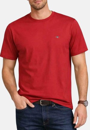 Uomo che indossa una maglietta rossa tinta unita a maniche corte, jeans blu scuro con cintura nera e orologio da polso marrone, in piedi con una mano in tasca.