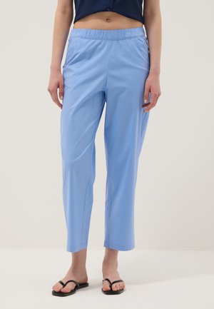 Mujer con pantalones recortados de cintura elástica color azul claro y chanclas negras, de pie sobre un fondo claro liso.