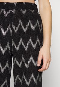 Czarne spodnie z srebrnym wzorem w zygzak. Materiał ma teksturowaną powierzchnię i średniej wysokości pas. Dłoń lekko opiera się na nodze.