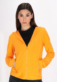 Felpa arancione con zip, dotata di cappuccio e due tasche laterali. Indossata sopra una maglietta nera, presenta una texture morbida.