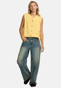 Gul strikket ermeløs topp med frontknapper, parret med løstsittende lyseblå jeans. Myk tekstur med en avslappet silhuett.