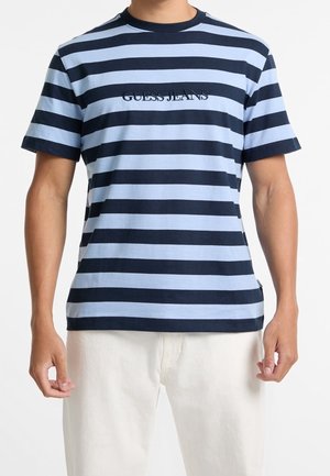 Hombre con camiseta de rayas horizontales azul marino y azul claro con texto "GUESS JEANS", combinada con pantalones color crema, de pie frente a un fondo liso.