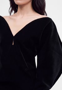 Femme portant une robe noire en velours épaules dénudées, avec de longues manches volumineuses et une petite fente à l'avant du col.