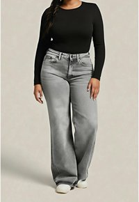 Femme portant un haut noir à manches longues, un jean gris taille haute à jambes larges et des chaussures blanches, debout sur un fond beige uni.