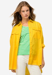 Camicia gialla con bottoni e due tasche sul petto, indossata sopra un top a canotta a righe verdi e bianche, abbinata a pantaloni bianchi. Tessuto morbido, vestibilità comoda.