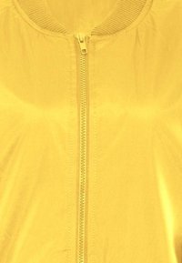 Giacca bomber gialla con zip frontale, colletto a coste e texture liscia. Il materiale è leggero e lucido, senza fantasie.