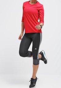 Czerwona sportowa koszulka z długim rękawem w zestawie z czarnymi legginsami o długości trzy czwarte z białymi paskami po bokach oraz czarnymi butami sportowymi.