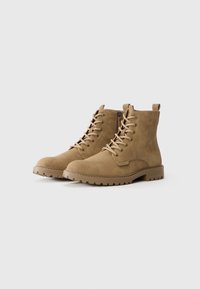 Par de botas de ante beige con cordones hasta el tobillo, con cremalleras laterales y suelas de goma robustas sobre un fondo blanco.