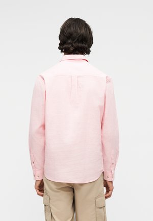 Man met donker haar draagt een lichtroze overhemd met lange mouwen en beige cargobroek, met de rug naar ons toe tegen een effen lichte achtergrond.