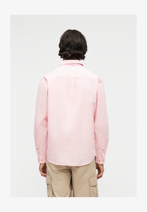 Man met donker haar draagt een lichtroze overhemd met lange mouwen en beige cargobroek, met de rug naar ons toe tegen een effen lichte achtergrond.