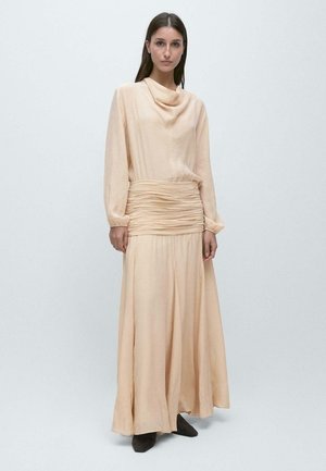 Kvinde med langt mørkt hår iført en lyse ferskenfarvet maxikjole med lange ærmer og en rynket taljebånd, samt mørke spidse sko, stående mod en ensfarvet baggrund.