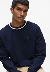 Sweatshirt bleu marine avec un col rond orné de rayures blanches et rouges. Logo discret sur la poitrine, texture douce, manches longues.