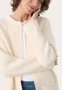 Cream textured knit cardigan with a round neck, loose fit, two front pockets, and a subtle pattern. Worn over a white shirt.