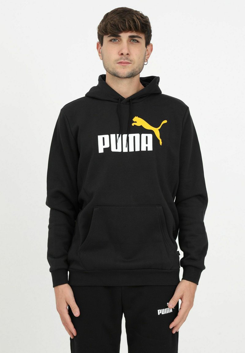 Sudadera negra de algodón con un bolsillo canguro, que presenta un logo blanco de "PUMA" y un gráfico de puma amarillo. Pantalones negros a juego visibles.