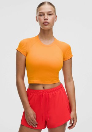 Mujer joven con una camiseta corta naranja de manga corta y pantalones cortos deportivos rojos, de pie frente a un fondo liso y claro.