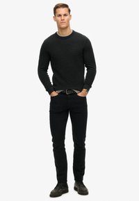 Svart, texturerad tröja med rund halsringning, kombinerad med slim fit svarta jeans och svarta läderskor. Modellen står med händerna i fickorna.