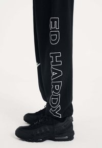 Schwarze Jogginghosen mit weißer "ED HARDY" Aufschrift, kombiniert mit schwarzen Sneakern, die ein strukturiertes Design und sichtbare Luftpolsterung aufweisen.
