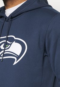 Felpa con cappuccio di colore blu navy realizzata in un tessuto morbido, dotata di un grande logo bianco dei Seahawks con dettagli verdi vicino all'occhio; include un cordino sul cappuccio.